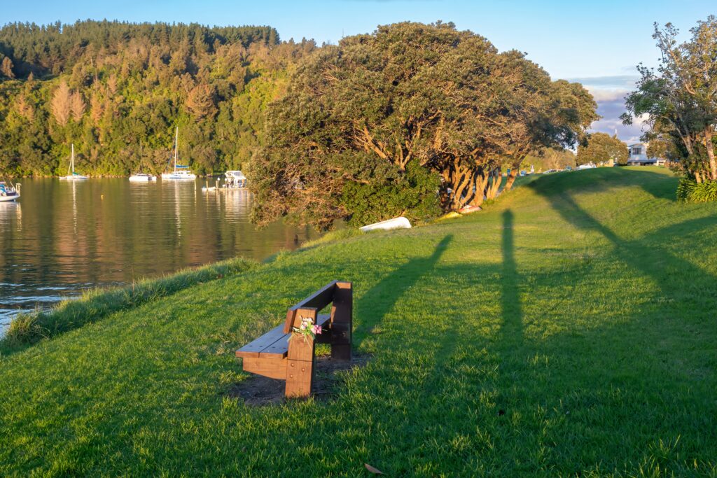 A wooden bench with flowers sits on a grassy bank shoreline overlooking a river with boats 